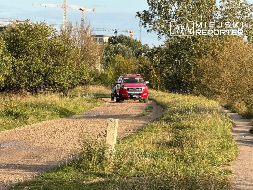 Czerwony samochód terenowy stoi na piaszczystej drodze w otoczeniu zieleni, z widocznymi dźwigami budowlanymi w tle.