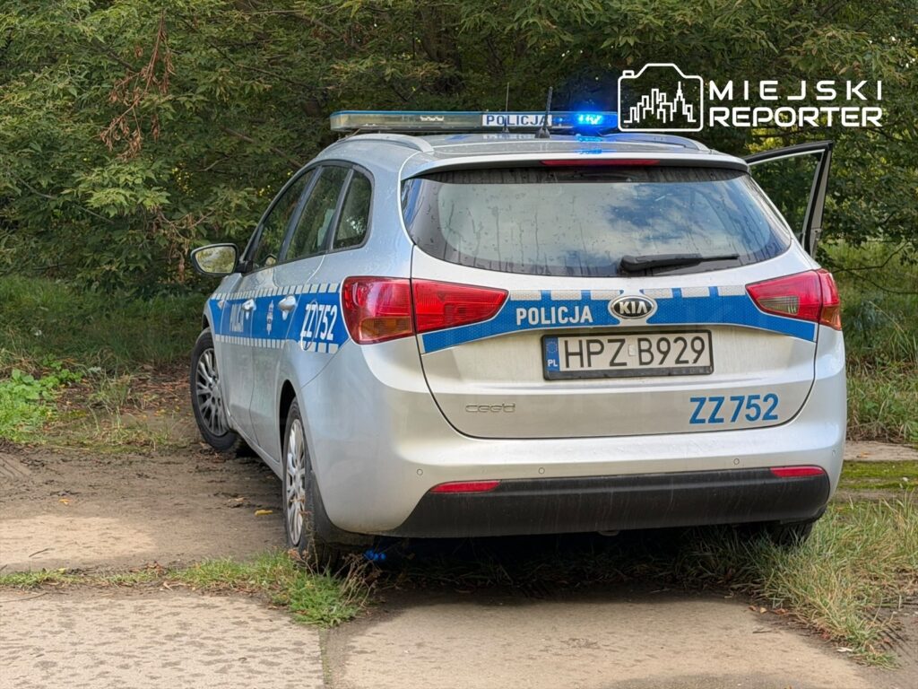 Policyjny samochód marki Kia z włączonymi sygnalizatorami stoi na poboczu drogi w otoczeniu zieleni.
