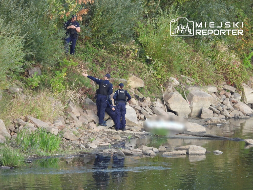 Policjanci prowadzą działania na brzegu rzeki, wskazując na miejsce, gdzie leży ciało.