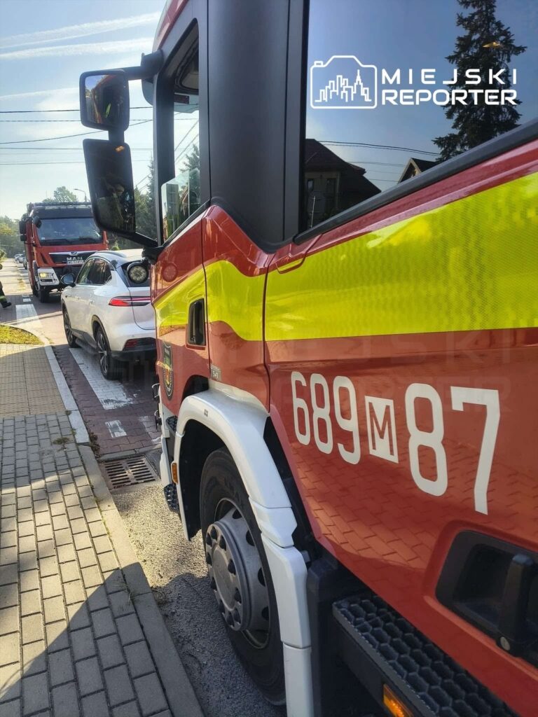 Czerwony wóz strażacki zaparkowany na ulicy, obok białego samochodu osobowego, w słoneczny dzień.