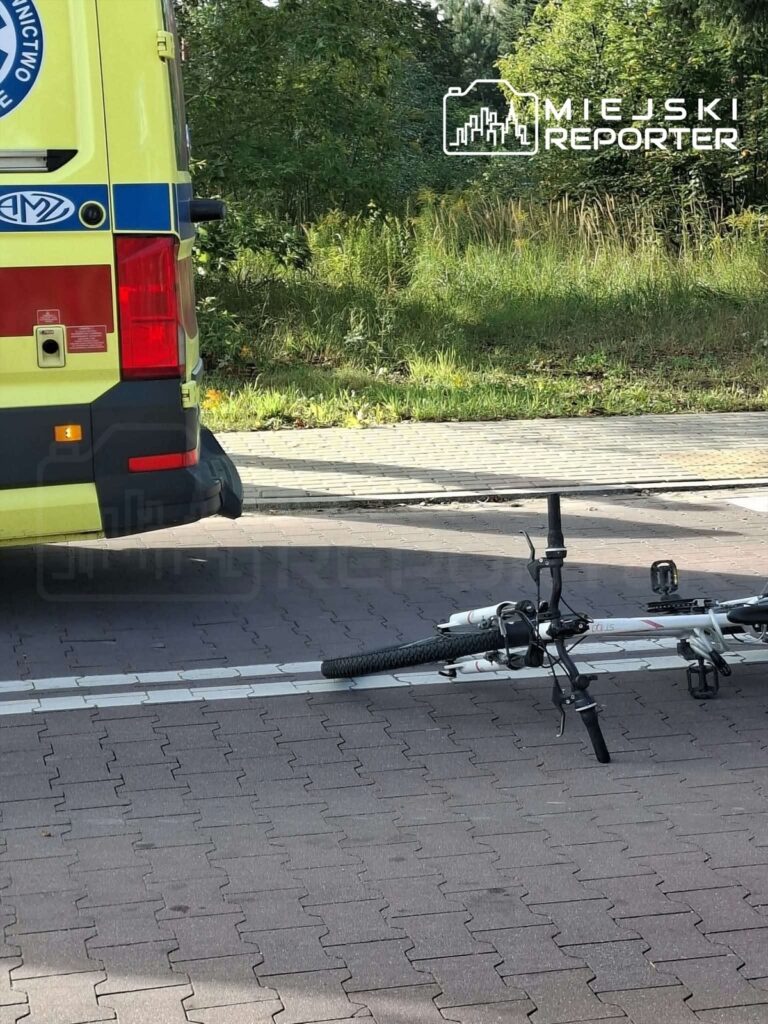 Na drodze leży uszkodzony rower obok karetki pogotowia, w tle zieleń drzew.