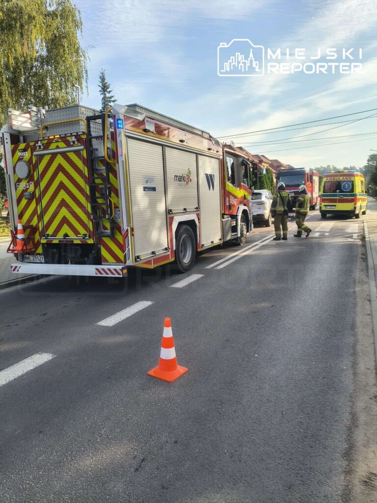 Wozidło strażackie z oznaczeniem "marki" stoi na drodze, obok dwóch strażaków w mundurach, a przed nim znajduje się pachołek ostrzegawczy.