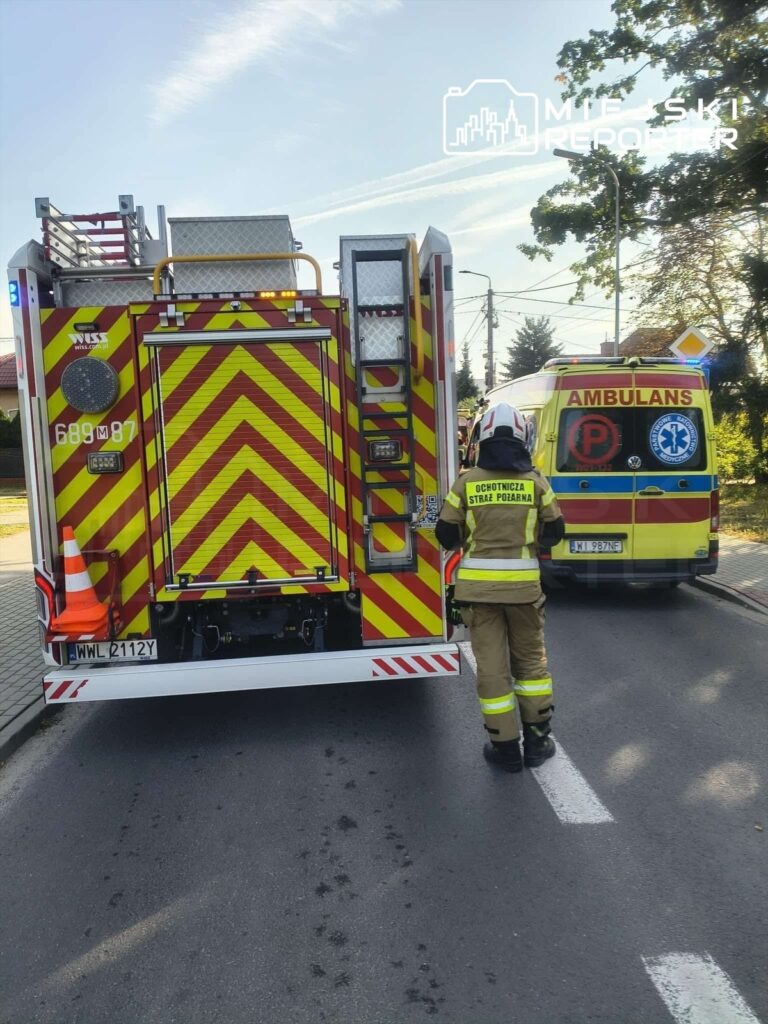 Strażak w mundurze podchodzi do wozu strażackiego na drodze, obok znajduje się ambulans.