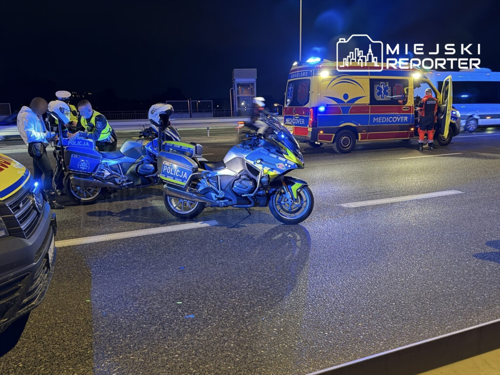 Policjanci na motocyklach rozmawiają z osobą w białej kurtce na drodze, obok stoi karetka z włączonymi sygnałami.