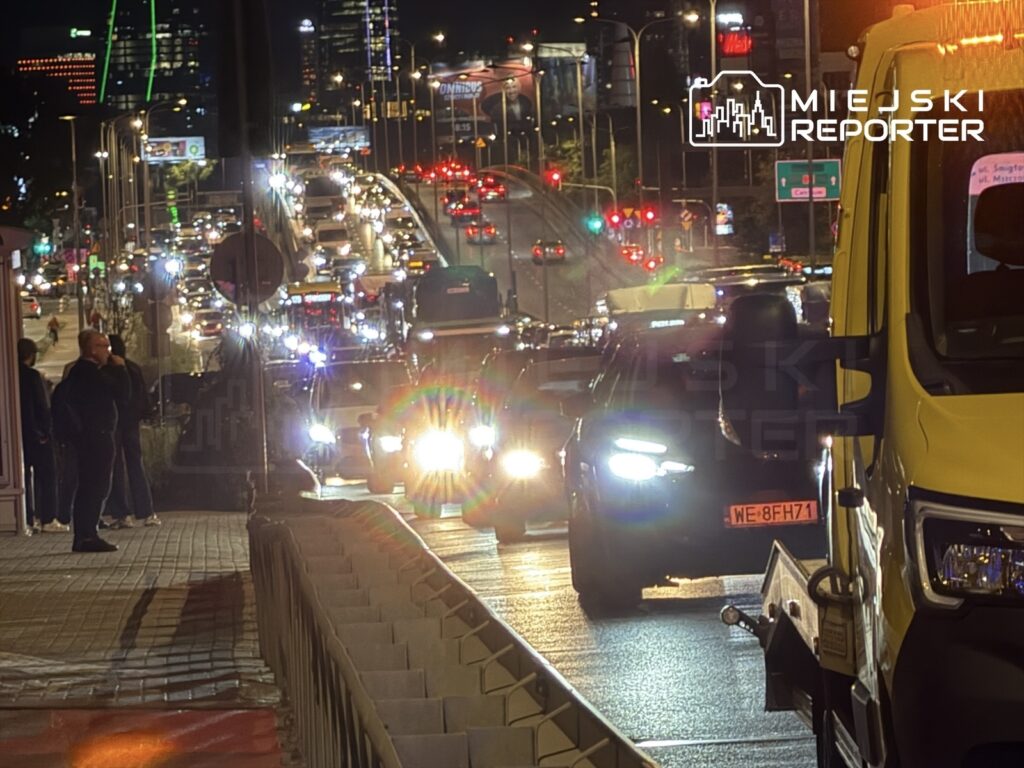Ruchliwe Aleje Jerozolimskie nocą, z samochodami stojącymi w korku i osobą czekającą na chodniku.