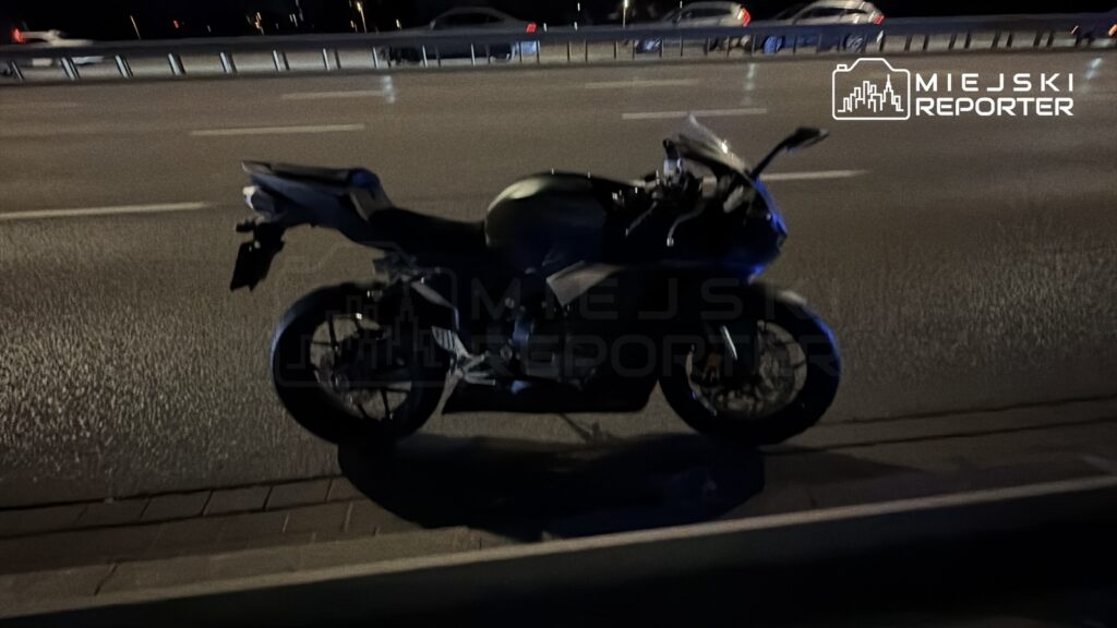 Czarny motocykl stojący na poboczu drogi w nocy, obok ruchliwej jezdni.