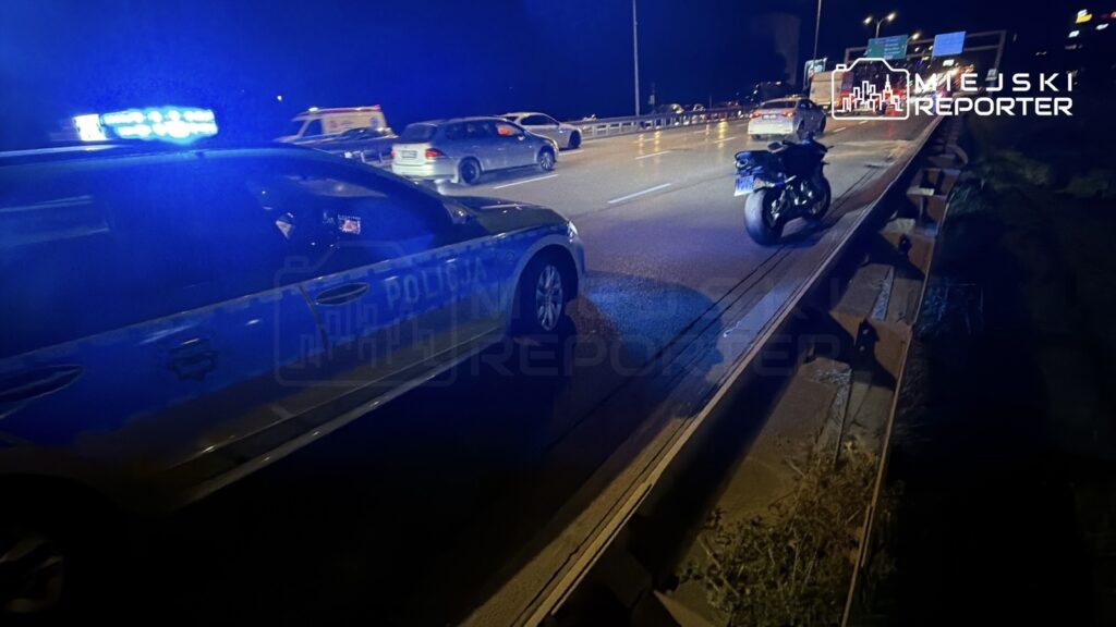 Policyjny radiowóz z niebieskimi światłami stojący na pasie drogi obok motocykla, w tle ruch samochodowy nocą.