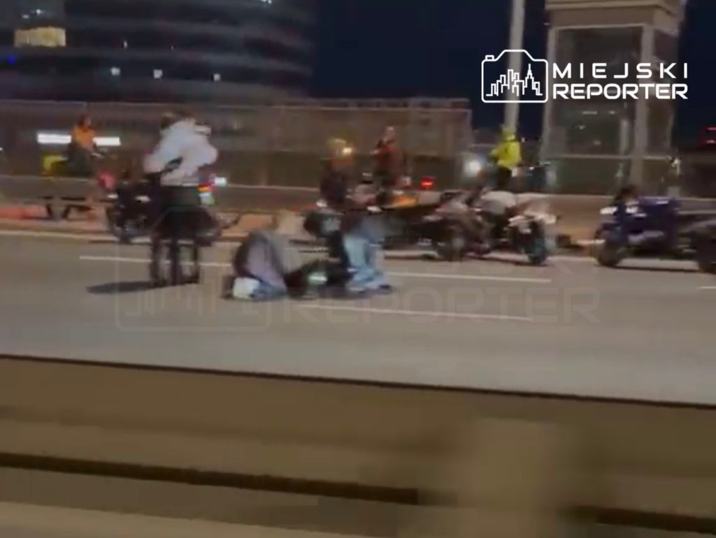 Grupa osób udziela pomocy poszkodowanym motocyklistom na jezdni w nocy, w tle widoczne motocykle.