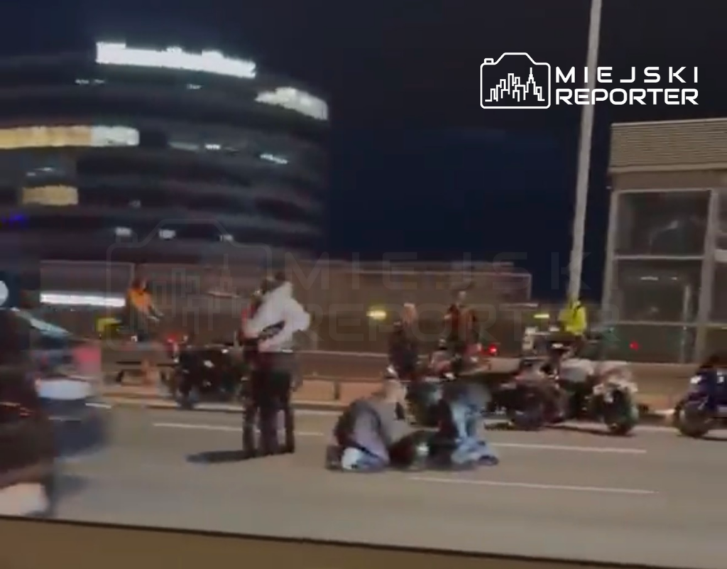 Grupa osób w kaskach otacza leżącego motocyklistę na drodze, obok stoją inne motocykle. W tle nowoczesny budynek.