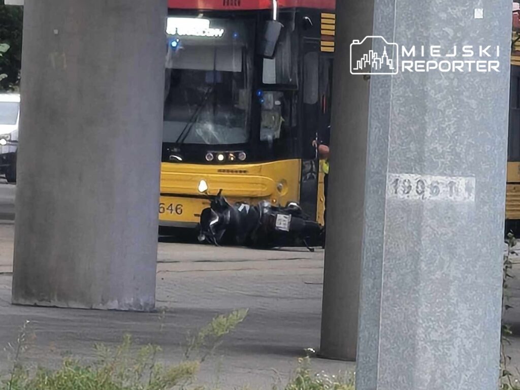 Motocyklista leży na ziemi obok tramwaju, w otoczeniu miejskich ulic i betonowych słupów.