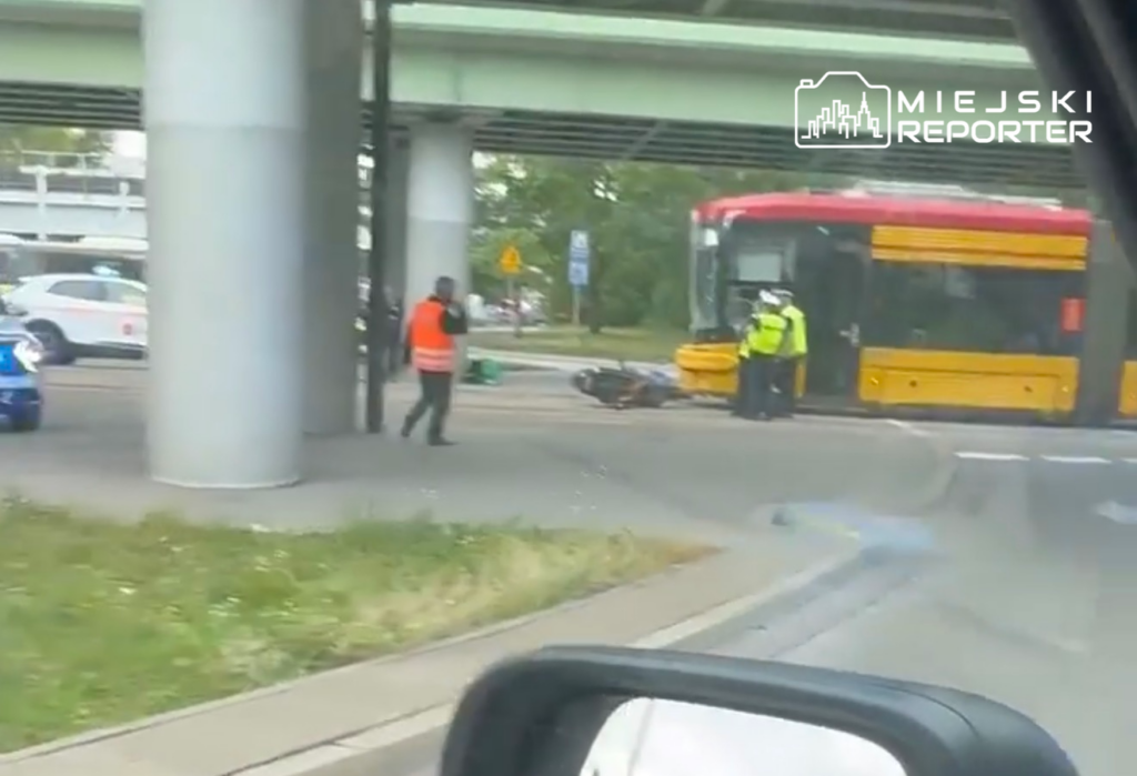 Motocyklista leży na jezdni obok zatrzymanego tramwaju, a obok interweniują policjanci w odblaskowych kamizelkach.