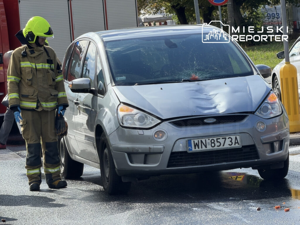 Strażak w hełmie i mundurze zbliża się do uszkodzonego samochodu osobowego po potrąceniu.