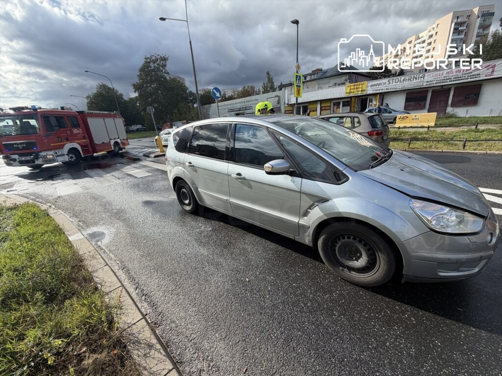 Na skrzyżowaniu stoi srebrny samochód osobowy, obok widać czerwony wóz strażacki. W tle widoczne są budynki mieszkalne.