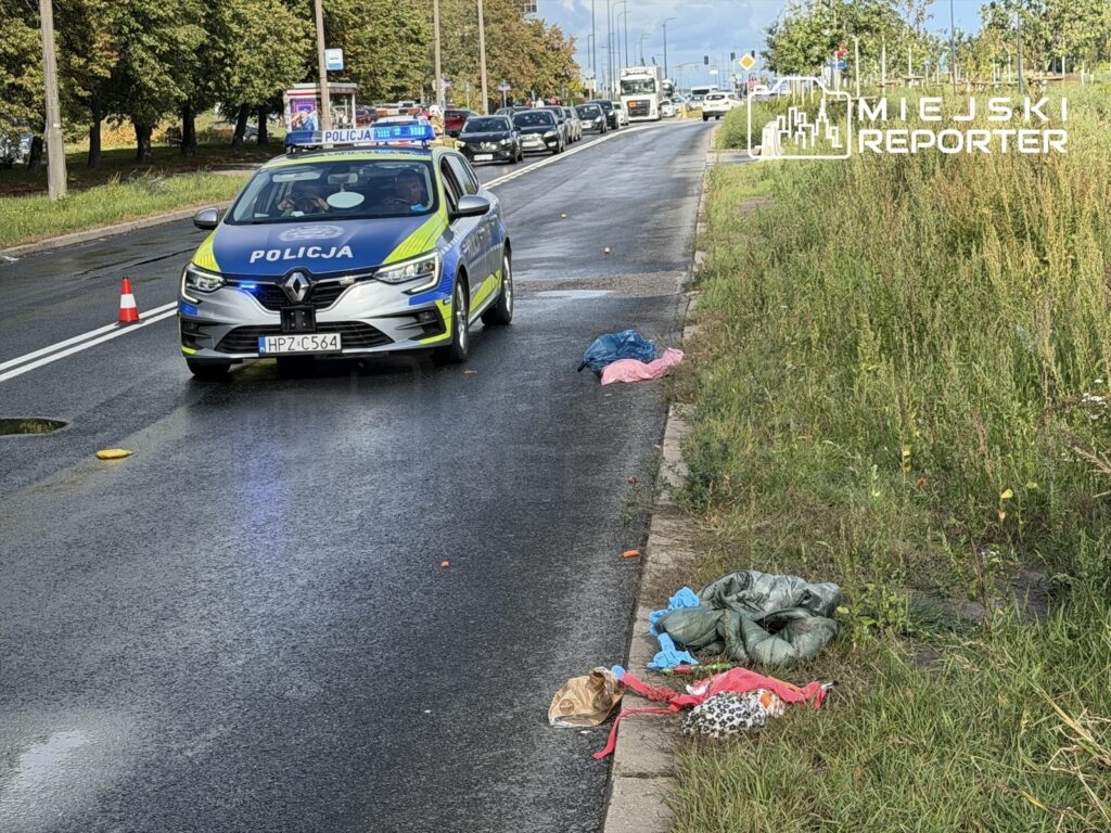 Policyjny radiowóz z włączonymi sygnałami świetlnymi zatrzymany obok jezdni, w pobliżu porzuconych ubrań po potrąceniu.