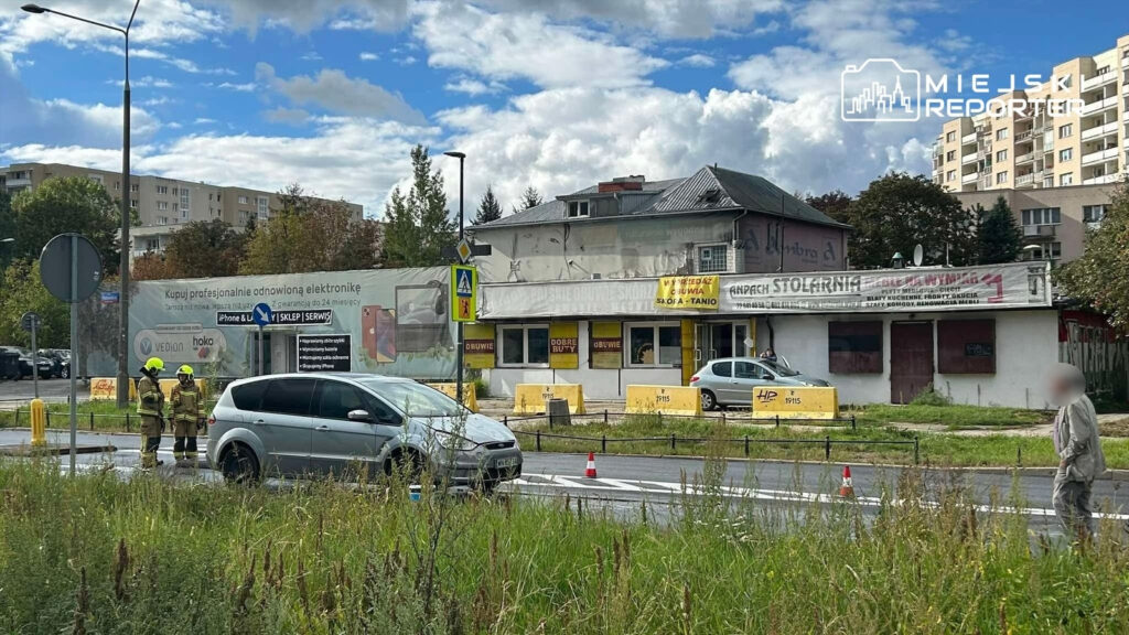 Na jezdni zatrzymany samochód osobowy, obok dwaj strażacy w mundurach, w tle budynki mieszkalne i reklamy. Potrącenie.