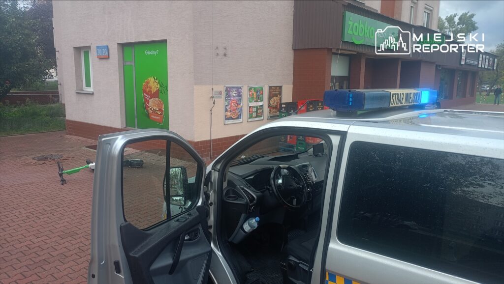 W tle widoczny zielony budynek z reklamą fast food, a z przodu otwarte drzwi radiowozu straży miejskiej. Na chodniku leży hulajnoga.