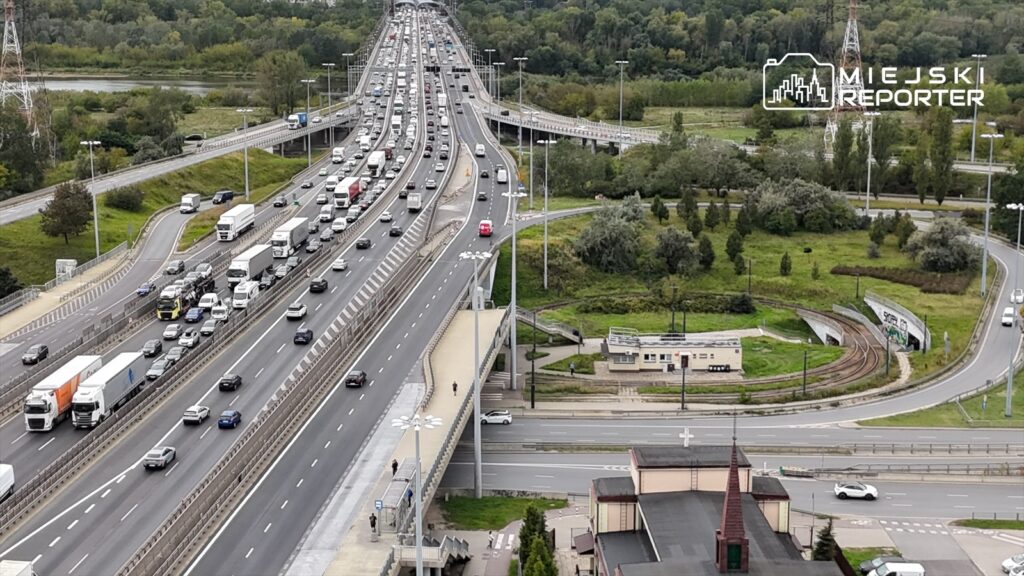 Ruchliwa trasa S8 z samochodami osobowymi i ciężarowymi, w tle zieleń i budynki w otoczeniu.