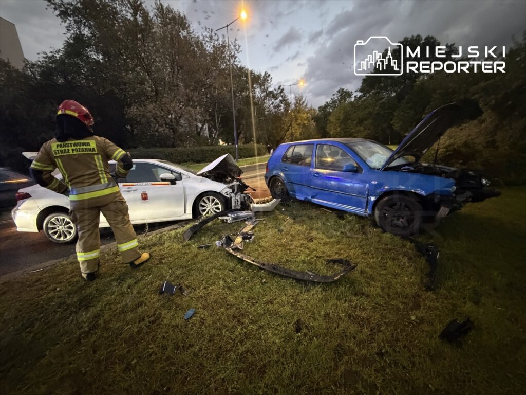 Strażak w mundurze obserwuje skutki wypadku drogowego z udziałem białego i niebieskiego samochodu.