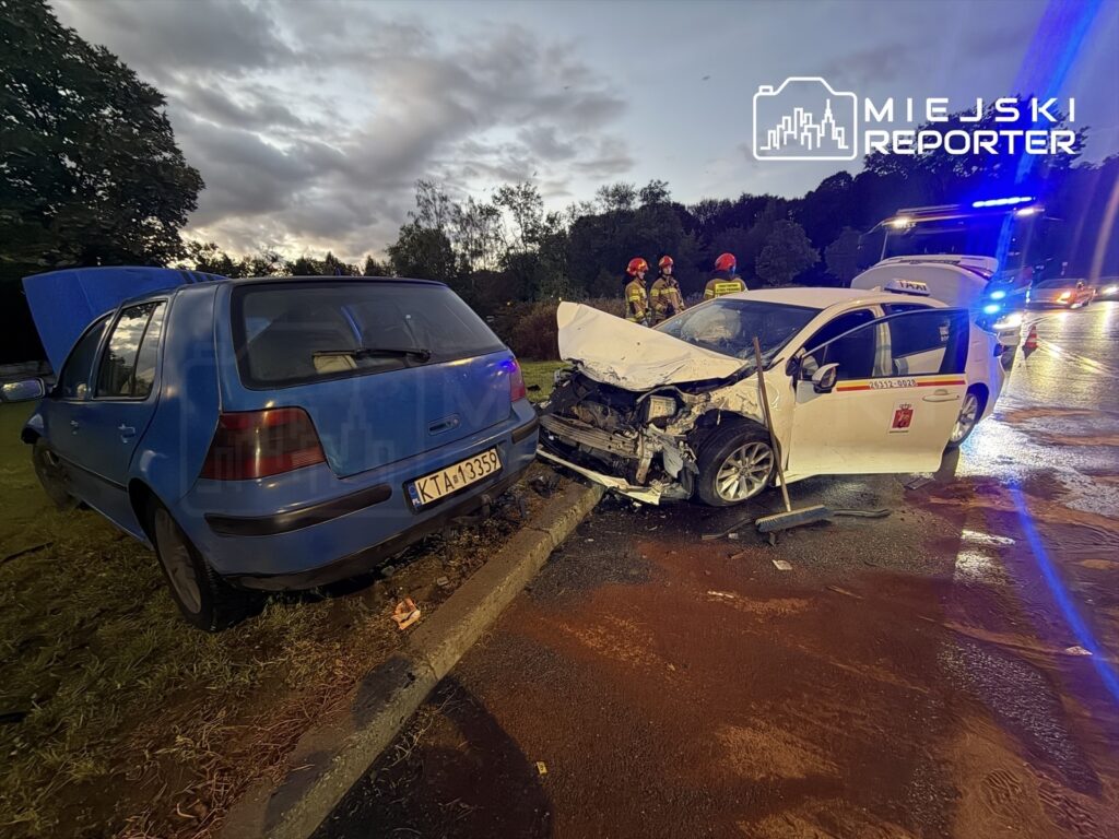 Dwa uszkodzone samochody, niebieski hatchback z otwartą maską oraz biały pojazd po kolizji, w otoczeniu służb ratunkowych.
