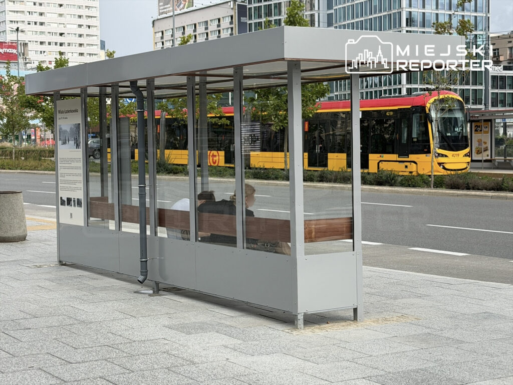 Nowoczesna wiata przystankowa z przeszkloną fasadą, obok zatrzymujący się tramwaj w miejskim otoczeniu.