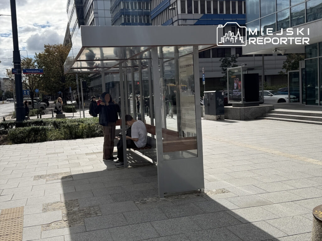 Mężczyzna siedzący na ławce w nowoczesnej wiacie, obok niego stoi inny mężczyzna, w tle przechodnie i samochody.