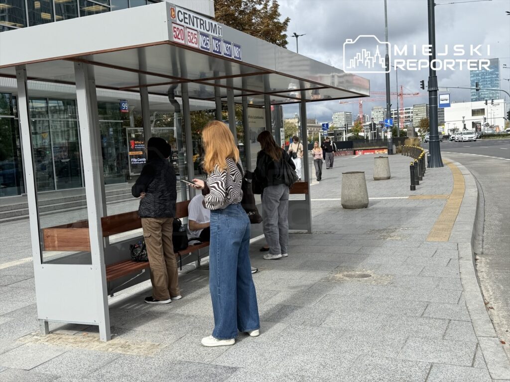 Grupa osób czeka na przystanku autobusowym z odrestaurowaną wiatą w centrum miasta.