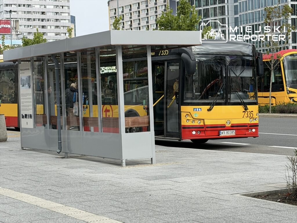 Nowoczesna wiata przystankowa z przeszklonymi ścianami stoi na chodniku, obok zatrzymującego się autobusu miejskiego.