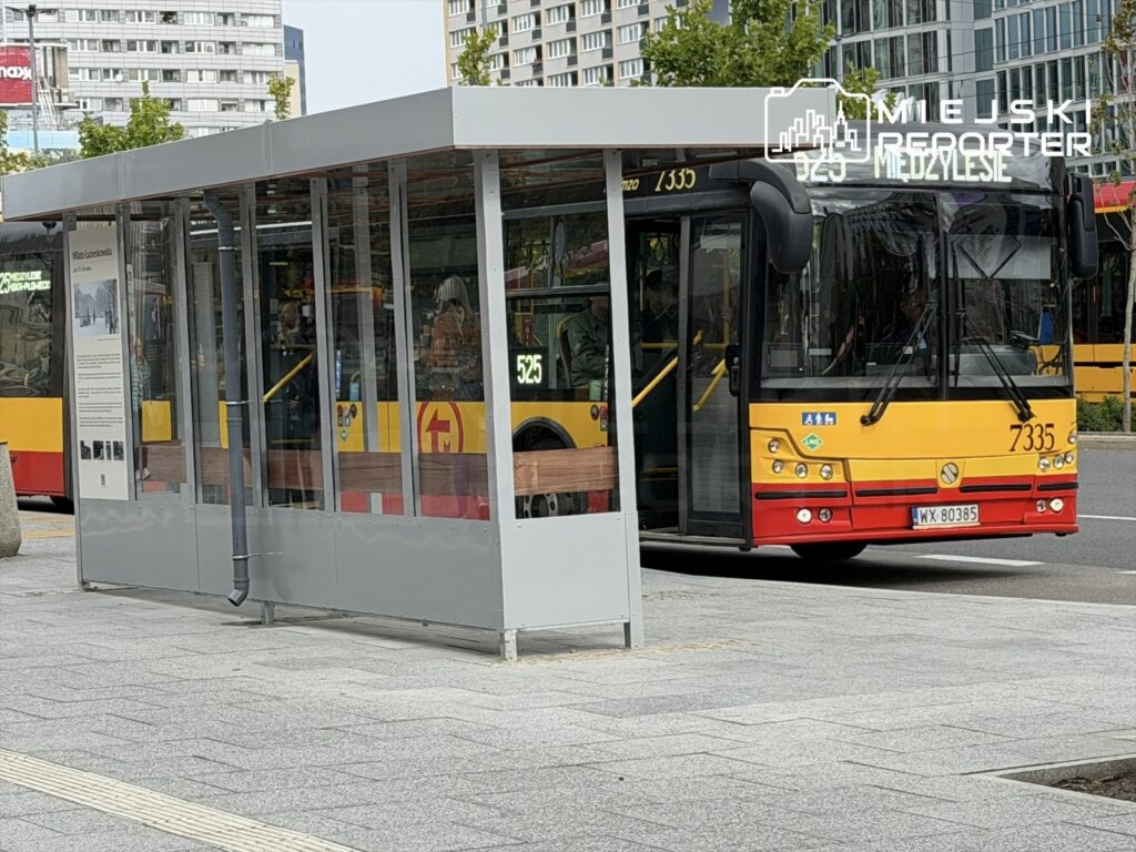 Nowa wiata przystankowa z przeszklonymi ścianami stoi na chodniku, obok nadjeżdżającego autobusu miejskiego.