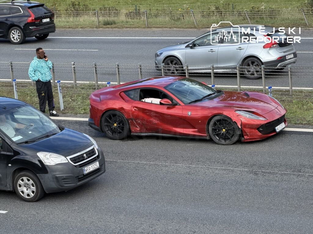 Mężczyzna w niebieskiej kurtce rozmawia przez telefon obok uszkodzonego czerwonego Ferrari na drodze ekspresowej.