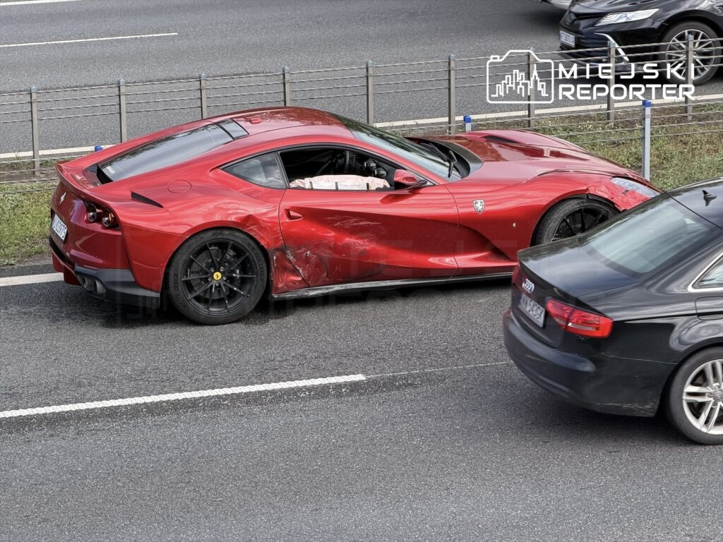 Czerwone Ferrari z uszkodzeniami na lewym boku stoi na pasie ruchu obok innego samochodu.