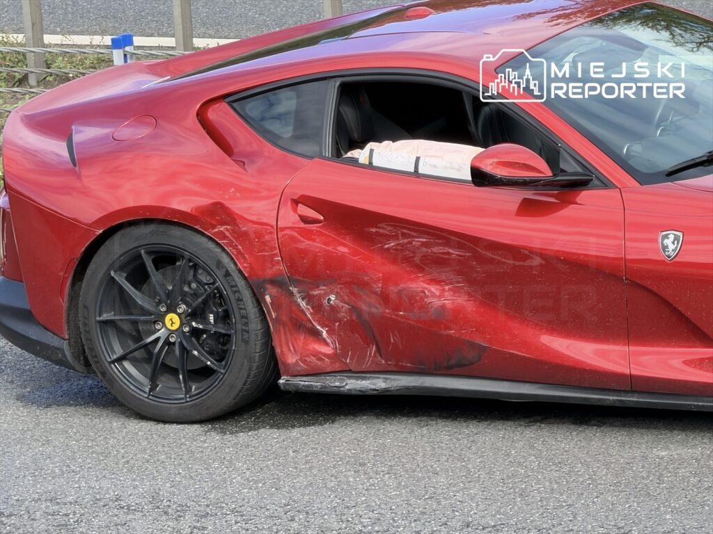 Czerwone Ferrari z uszkodzoną boczną częścią stoi na drodze, obok widoczny jest asfalt i barierka ochronna.