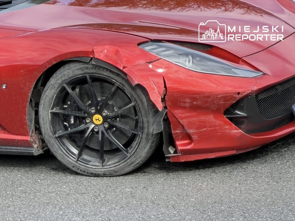 Czerwone Ferrari z uszkodzoną przednią częścią i zniszczonym kołem stojące na asfalcie.