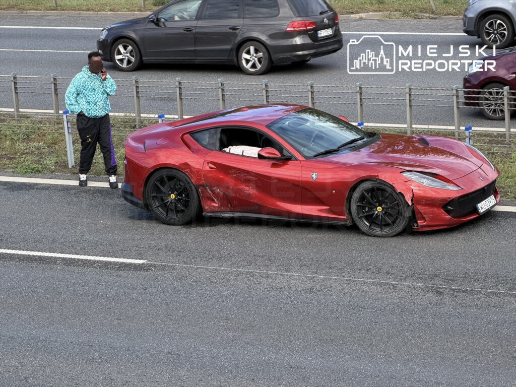 Mężczyzna w niebieskiej kurtce rozmawia przez telefon obok uszkodzonego czerwonego Ferrari na drodze.