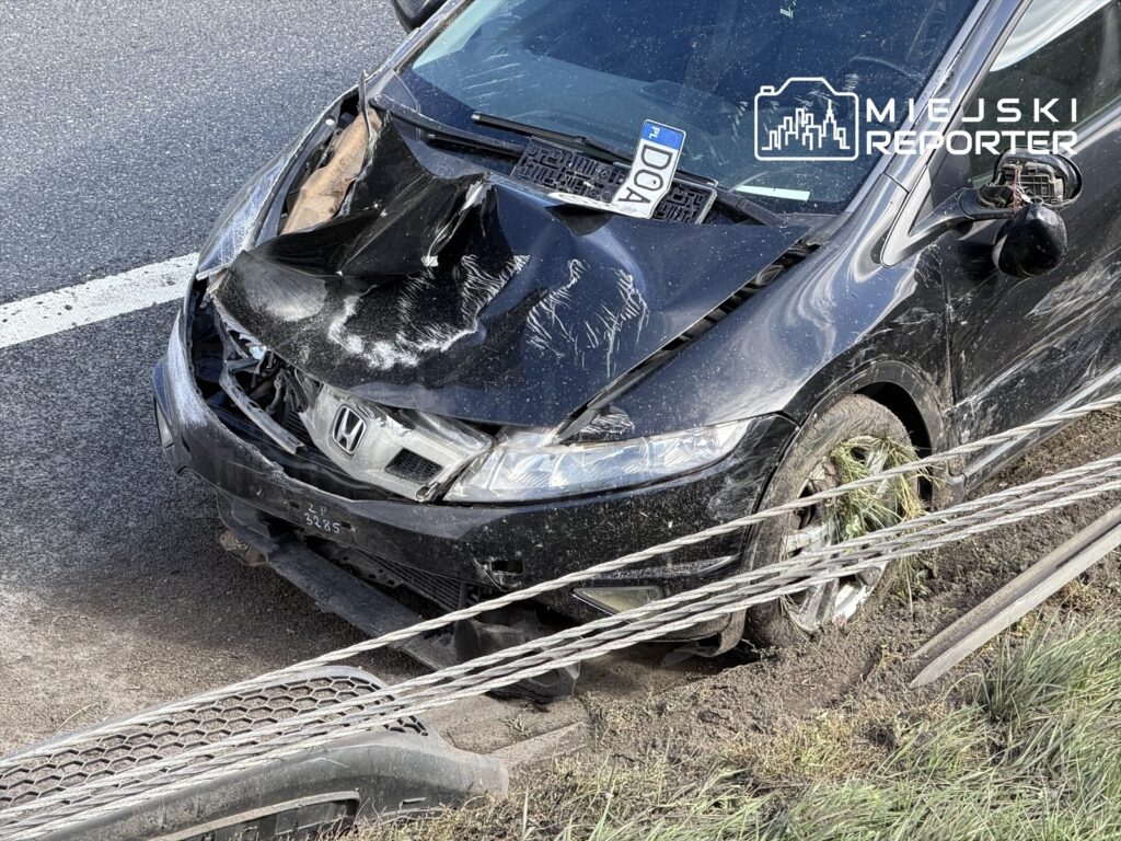 Zniszczony czarny samochód osobowy marki Honda z uszkodzoną przednią częścią stoi na poboczu drogi, obok ogrodzenia.