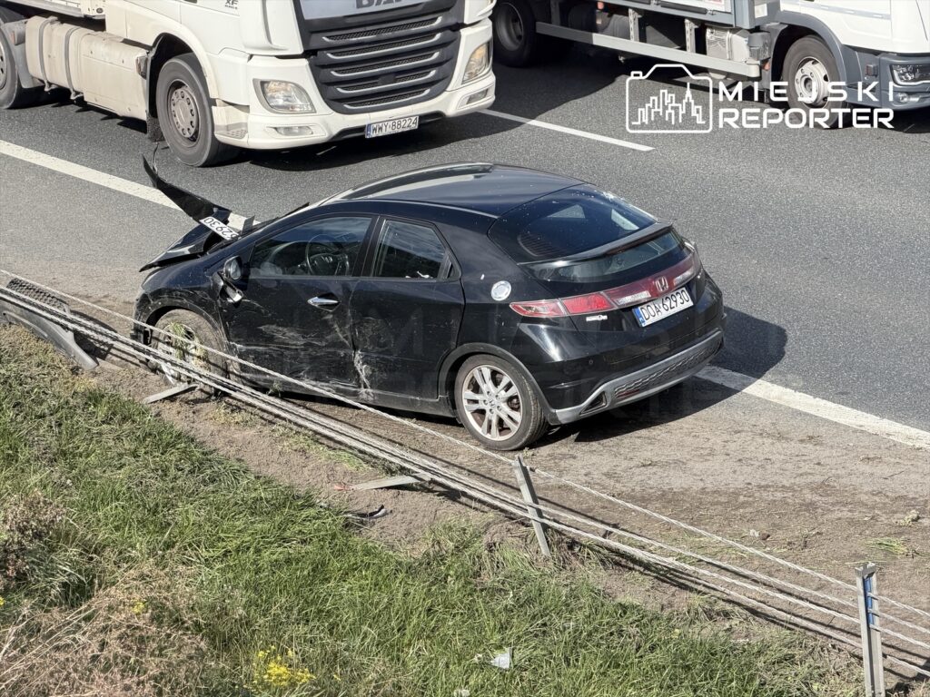 Czarny samochód osobowy z uszkodzeniami stoi na poboczu drogi, obok przejeżdżających ciężarówek.