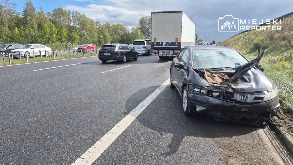 Zniszczony czarny samochód osobowy z otwartą maską stoi na poboczu drogi, obok przejeżdżających pojazdów.
