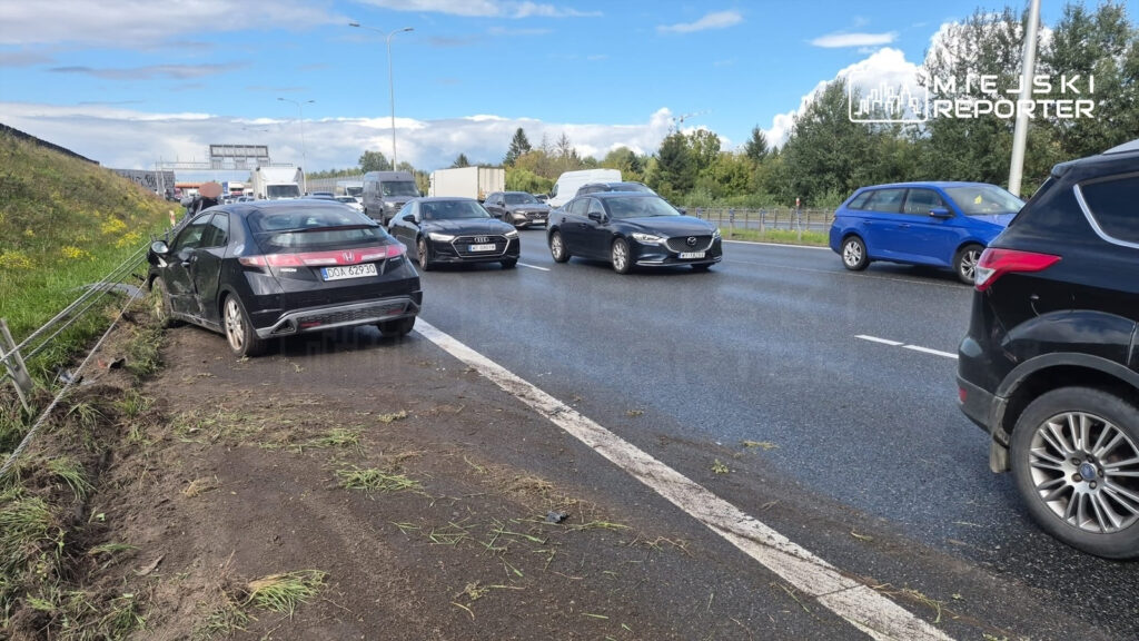 Czarny samochód osobowy stoi na poboczu drogi, z uszkodzoną przednią częścią, obok ruchliwej jezdni z innymi pojazdami.