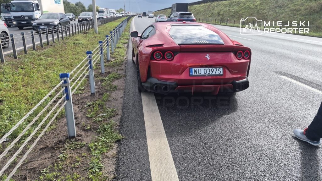 Czerwone Ferrari stoi na poboczu drogi, obok ruchliwej trasy, z widocznymi samochodami w tle.