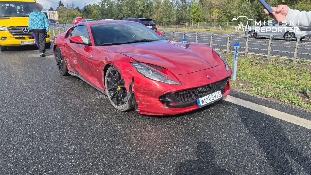 Uszkodzone czerwone Ferrari z zniszczonym przodem stoi na drodze, obok mężczyzna z telefonem w ręku.