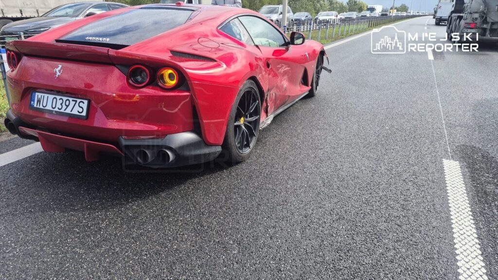 Uszkodzone czerwone Ferrari stoi na pasie ruchu, z widocznymi zniszczeniami bocznej części nadwozia.