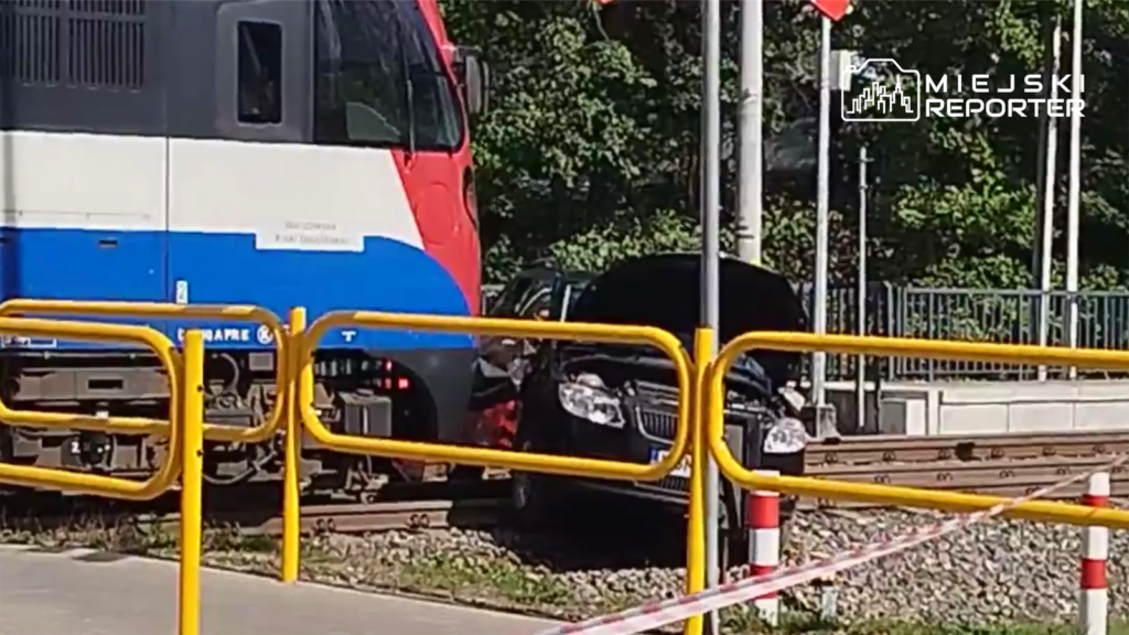 Pociąg pasażerski zbliża się do uszkodzonego samochodu zaparkowanego na torach kolejowych, otoczonego barierkami.
