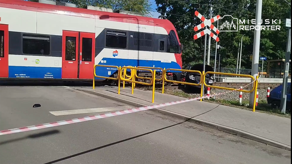 Pociąg pasażerski zatrzymany przy przejeździe kolejowym, otoczony żółtymi barierkami i taśmą ostrzegawczą.