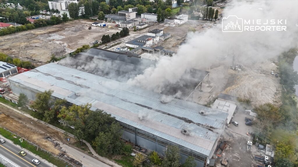 Hala przemysłowa z dymem unoszącym się z dachu, otoczona placem budowy i drzewami, w pobliżu ulicy Płochocińskiej.