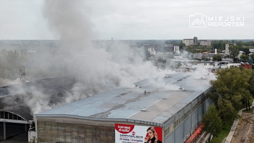Gęsty dym unoszący się z dachu hali przemysłowej, otoczonej zielenią i innymi budynkami, przy ulicy Płochocińskiej.