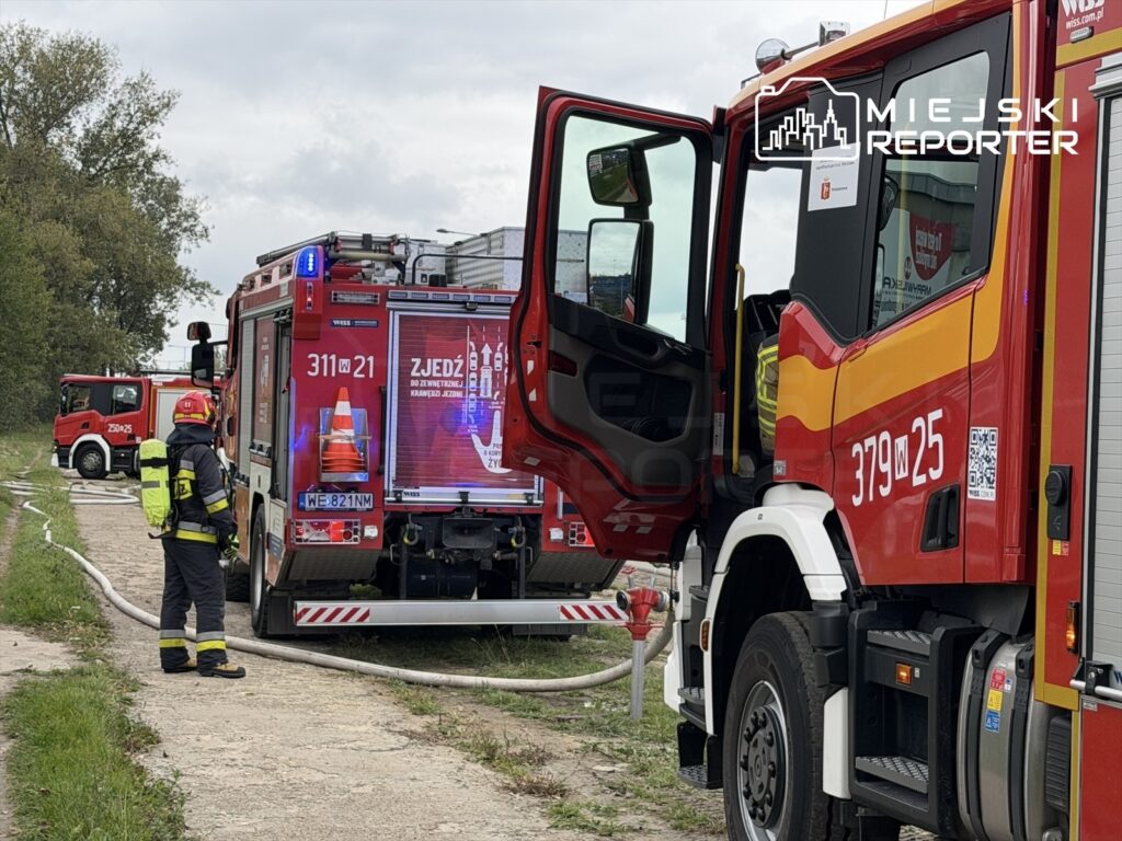 Strażak w mundurze przy wozie strażackim z otwartymi drzwiami, w tle inne pojazdy gaśnicze na ulicy Płochocińskiej.