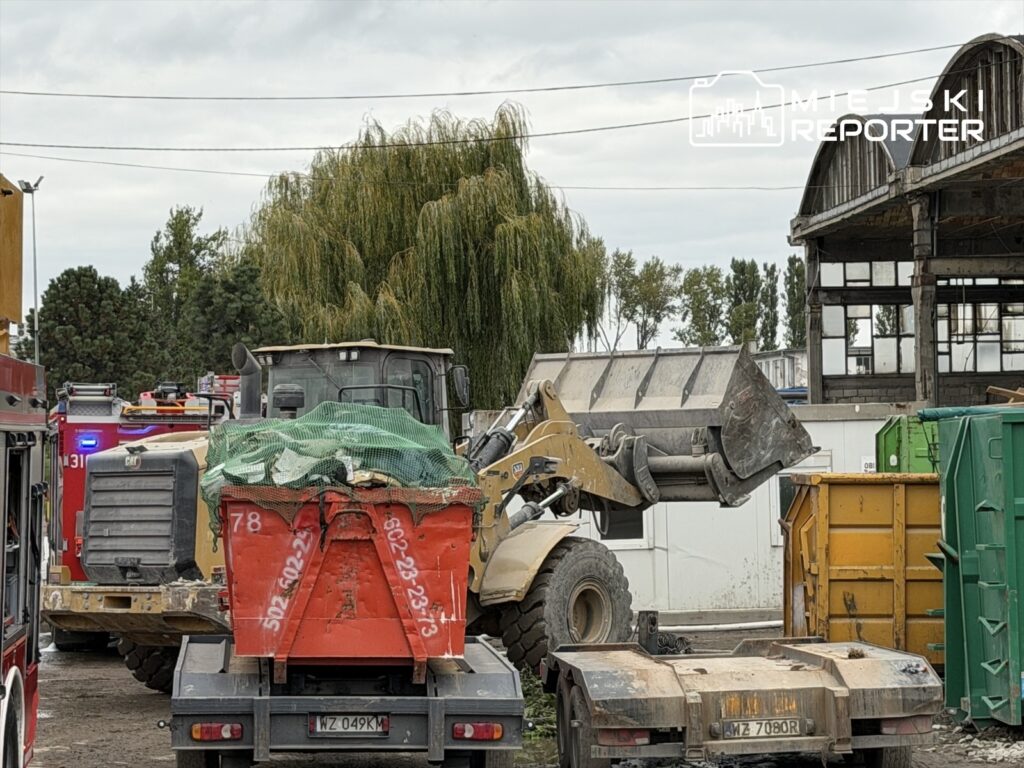 Ciężki sprzęt budowlany podnosi kontener na placu, w tle widać straż pożarną i zniszczony budynek. Akcja trwa przy Płochocińskiej.