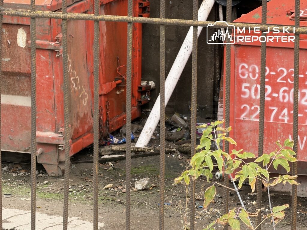 Czerwone kontenery na odpady umieszczone za metalowym ogrodzeniem, otoczone zanieczyszczeniami i chwastami.