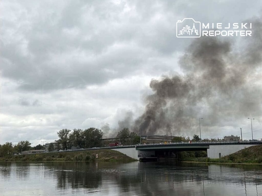 Czarny dym unoszący się nad budynkiem w tle, widoczny z perspektywy rzeki, pod chmurami.