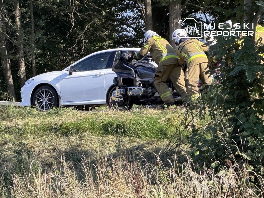 Strażacy transportują uszkodzony motocykl obok białego samochodu na poboczu drogi otoczonej drzewami.