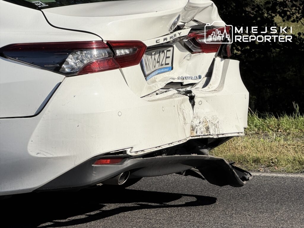 Uszkodzona biała Toyota Camry z wgniecionym tyłem, stojąca na poboczu drogi, z widocznymi zarysowaniami i uszkodzeniami.